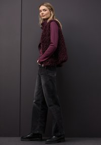 Bordeauxfarbene Fleeceweste mit strukturierter Stoffoberfläche, getragen über einem eng anliegenden Langarmshirt, kombiniert mit schwarzen Jeans mit weitem Bein und schwarzen Stiefeletten.