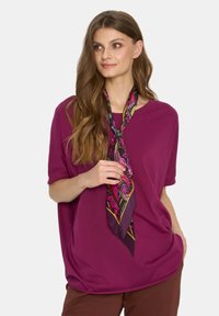 Luźna fioletowa koszulka z miękkiego materiału, w poł ączeniu z kolorowym jedwabnym szalikiem związanym wokół szyi. Brązowe spodnie.