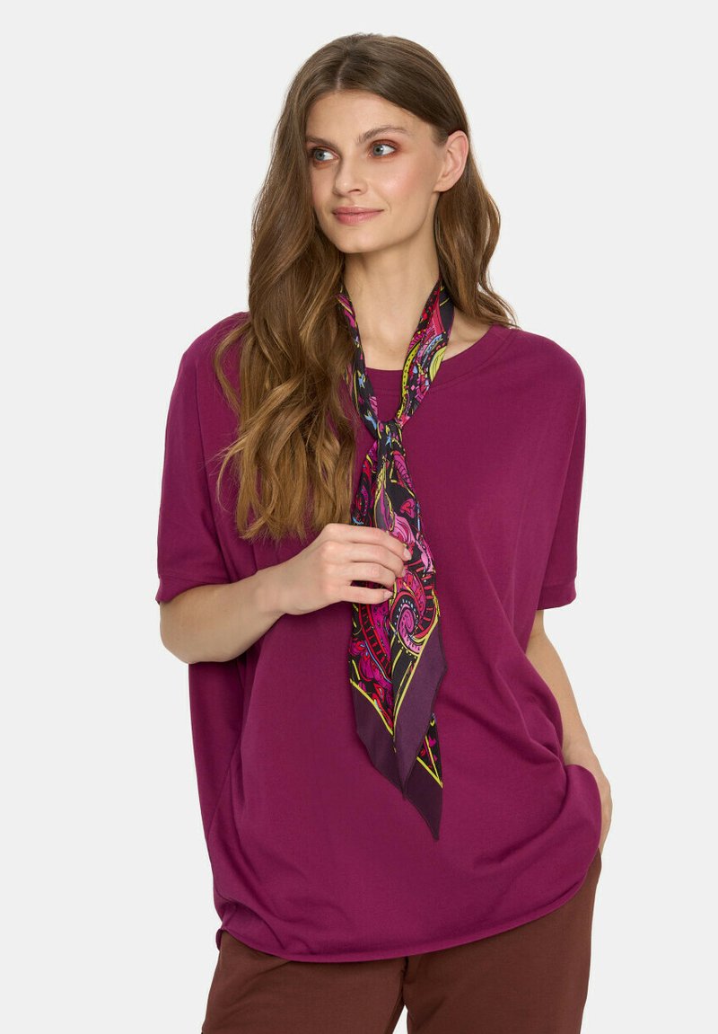 Luźna fioletowa koszulka z miękkiego materiału, w połączeniu z kolorowym jedwabnym szalikiem związanym wokół szyi. Brązowe spodnie.