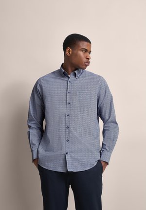 Blause Hemd mit Muster, mit Kragen zum Knöpfen, langen Ärmeln und einer Tasche auf der linken Seite; kombiniert mit dunklen Hosen.