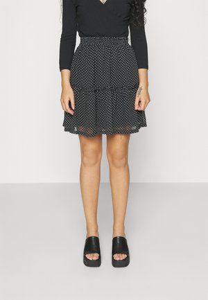 Woman wears black long-sleeve top, black skirt with white polka dots, and black platform open-toe shoes, standing against white background.
