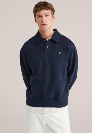 Jonge man met een marineblauwe lange mouw trui met kraag en witte broek, staande tegen een effen beige achtergrond.