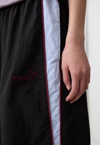 Un gros plan d'une main reposant sur un pantalon noir avec une rayure latérale bleue et bordeaux, avec "FENTY" brodé en bordeaux.