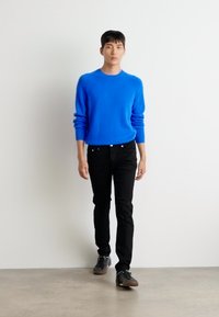 Jersey de punto azul con mangas largas, combinado con pantalones ajustados negros y zapatos oscuros, presentado sobre un fondo blanco liso.