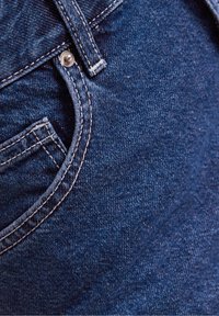 Tmavě proplálená džínovina s viditelnou kapsou a stříbrným knoflíkem. Kontrastní prošívání podél švů a okrajů kapes.