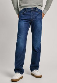 Vaqueros oscurs de mezclilla, con un corte recto clásico, diseño de cinco bolsillos y costuras de cobre. Combinados con zapatillas blancas.