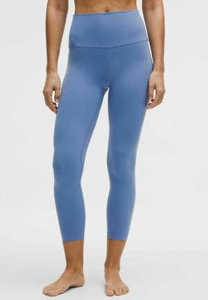 Kobieta ubrana w wysokie, sięgające kostek niebieskie legginsy, stojąca boso na jednolitym białym tle.