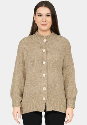 Femme aux longs cheveux bruns portant un cardigan en grosse maille beige avec six gros boutons, debout devant un fond blanc uni.