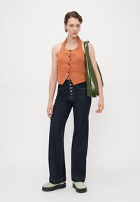 Oransje avkortet halter-top med knapper, kombinert med mørkeblå flare-jeans og grønne platå-sandaler. Modellen bærer en blank grønn tote-bag.