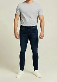 Vaqueros ajustados azul marino con diseño de cinco bolsillos, combinados con una camiseta de manga corta gris claro y zapatos casuales blancos.