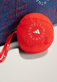 Bolso rojo plegable con forma redonda, textura suave y logotipo blanco. Incluye una correa de textil con marca roja.