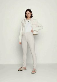 Casaco cropped bege claro com dois bolsos no peito, combinado com jeans skinny bege de cintura alta e sandálias taupe. Top branco por baixo.