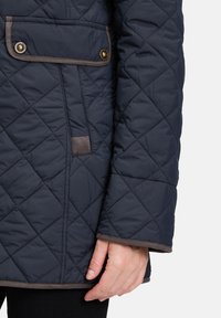 Marineblå quilted jakke med brune accenter, der har en lomme med trykknap og en lige skæring. Foringen er glat og velegnet til køligere vejr.
