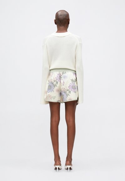 Suéter blanco de punto, mangas largas, y pantalones cortos florales crema con cinturilla verde. Modelo con tacones blancos, posando de espaldas contra un fondo claro.
