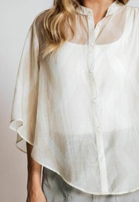 Sheer cream blouse with a button front, short sleeves, and a flowing silhouette. Features a subtle geometric pattern and a curved hemline.