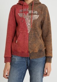 Bluza z kapturem o podzielonym wzorze, jedna połowa czerwona, druga brązowa, z logo graficznym i sznureczkami do ściągania. Materiał to mieszanka bawełny o teksturowanym wyglądzie.