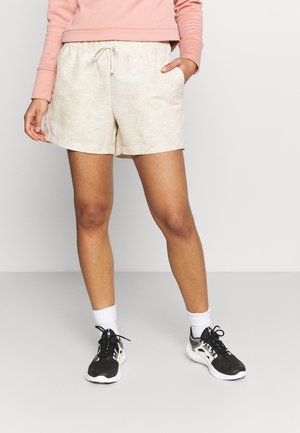 Personne portant un short beige clair avec cordon, un haut à manches longues rose, des chaussettes blanches et des chaussures de sport noir et blanc, debout devant un fond uni.
