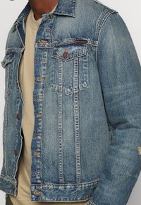 Jeansjacka med ljusblåtvätt, med knappstängningar, två bröstfickor och en urtvättad textur med slitna detaljer.