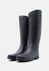 Ζευγάρι ψηλών μαύρων λαστιχένιων μπότες βροχής με ομαλή ματ επιφάνεια και ετικέτα με το λογότυπο στο πάνω μπροστινό μέρος, στέκονται πάνω σε λευκό φόντο.