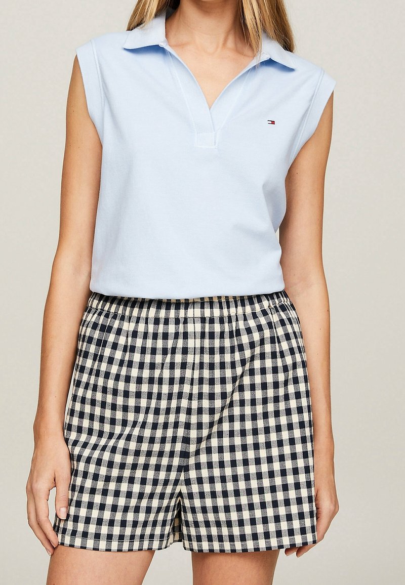 Polo sans manches bleu clair associé à un short à carreaux bleu marine et blanc taille haute sur une personne debout.