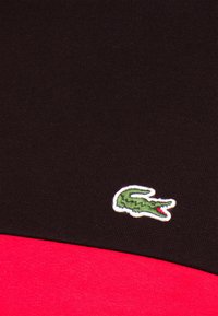 Svart och röd färgblockerad tyg, med en broderad grön krokodil-logotyp, slät textur och atletisk design med kontrasterande färger.