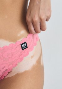 Pink lace thong with scalloped edges, featuring a black "HUGO" tag. The material is sheer, revealing skin underneath with light patterns.