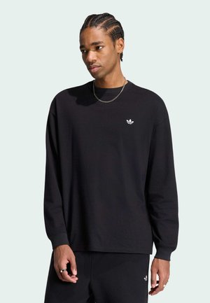 Ung mand med flettet hår iført en sort Adidas langærmet trøje og matchende bukser mod en ensartet lys baggrund.