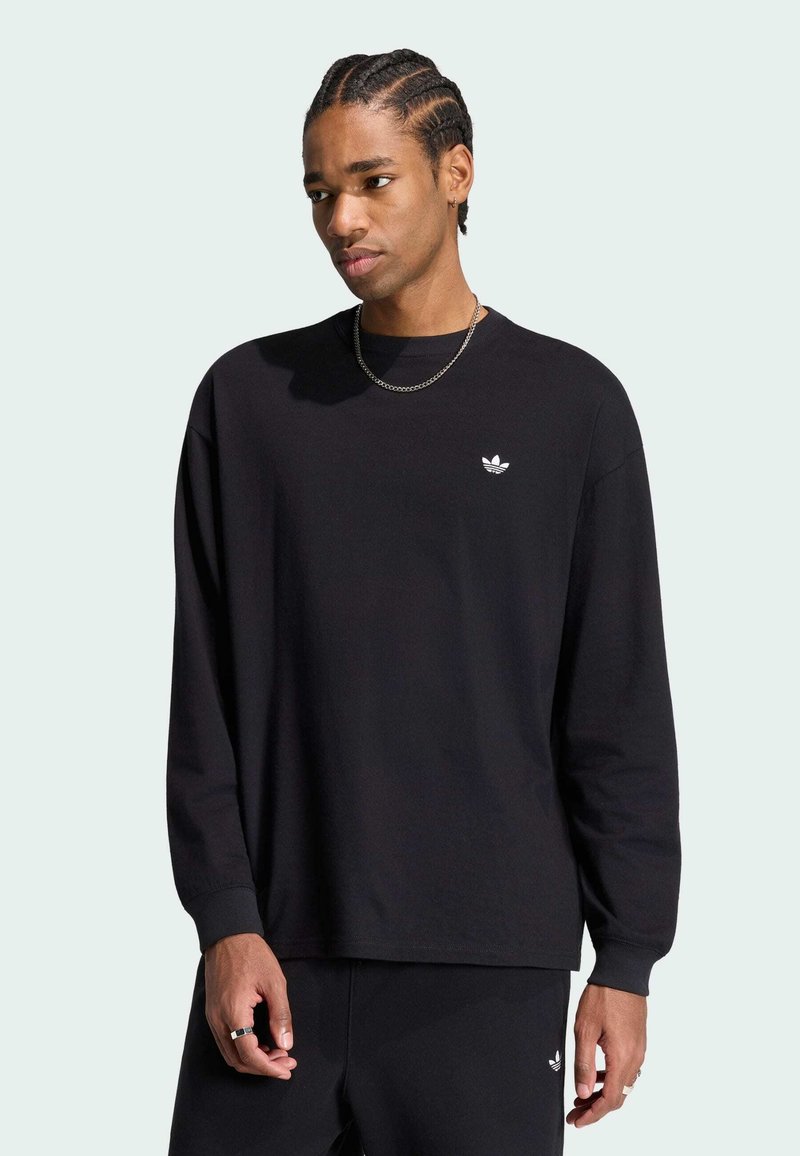 Jeune homme aux cheveux tressés portant un t-shirt noir Adidas à manches longues et un pantalon assorti sur un fond clair uni.