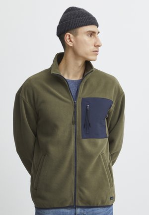 Fleece takki, joka on oliivinvihreä ja jossa on tummansininen rintatasku. Takissa on vetoketjukiinnitys ja sivutaskut, teksturoitu kangas sekä pystykaulus.