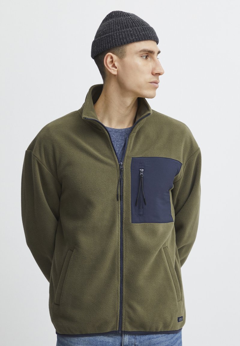 Fleece jaciņš ar rāvējslēdzēju olīvu zaļā krāsā ar tumši zilu krūšu kabatu, rāvējslēdzēja aizdare, sānu kabatām, teksturētu audumu un stāvošo apkakli.