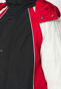 Sektion af en jakke med røde ærmer, hvide sidelapper og en sort front med trykknapper og høj krave.