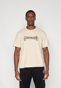 Μπεζ βαμβακερό μπλουζάκι με στρογγυλή λαιμόκοψη, κοντά μανίκια και μαύρο κείμενο "Grateful Dead" στο στήθος.