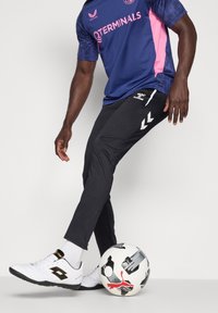 Oseba, oblečena v mornarsko modro in roza športno majico ter črne hlače, z desno nogo kontrolira belo žogo Puma.