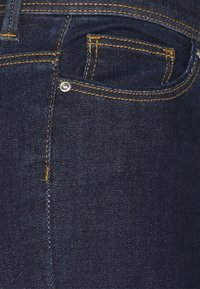 Tela de mezclilla azul oscuro con costuras naranjas detalladas y un pequeño remache en el bolsillo frontal, mostrando de cerca el área del bolsillo delantero de los vaqueros.