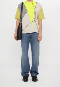 Homme portant un gilet tricoté géométrique jaune et beige par-dessus une chemise jaune à manches courtes, un jean bleu, des chaussures noires et portant un sac à bandoulière en cuir noir.