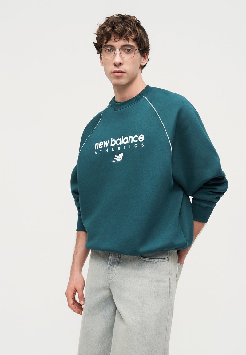 Jeune homme portant des lunettes, un sweat New Balance Athletics bleu sarcelle, et un jean gris clair, debout devant un fond blanc uni.