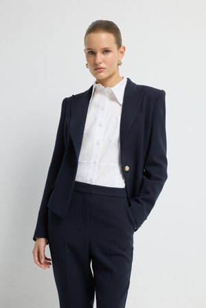 Blazer ajustado de color azul marino con hombreras, un único botón dorado, acompañado de una camisa blanca tipo botón y pantalones a juego en azul marino. Texturas suaves.