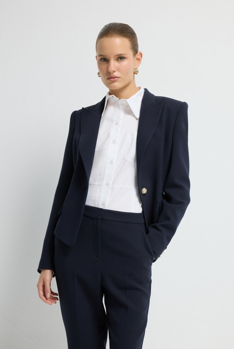 Navy sako na míru s vycpávkami na ramena, jedním zlatým knoflíkem, spárované s bílou košilí a ladícími námořnickými kalhotami. Hladké textury.