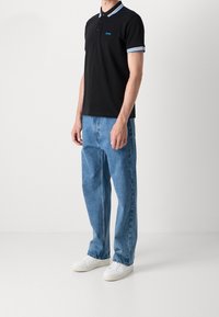 Homme portant un polo noir avec des bordures bleu et blanc, un jean bleu clair et des baskets blanches, se tenant devant un fond uni.