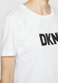 Vit bomull t-shirt med korta ärmar. Har en stor svart broderad "DKNY" logotyp på framsidan, rund halsringning och mjuk textur.