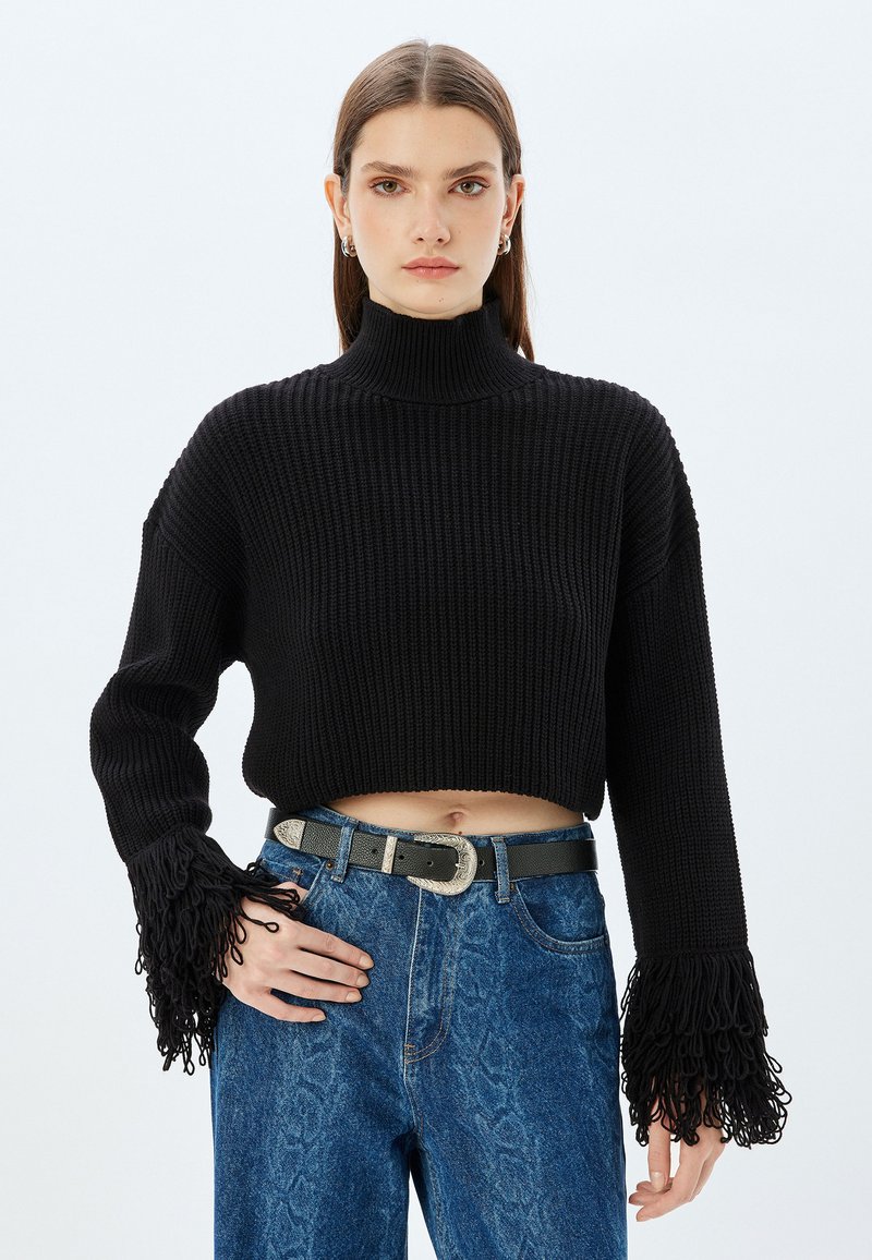 Schwarzer gestrickter, verkürzter Pullover mit hohem Kragen und langen Ärmeln, der mit Fransenakzenten an den Manschetten versehen ist, getragen mit hochgeschnittenen blauen Jeans.
