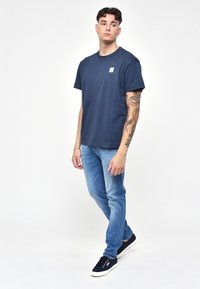 Námořnickomodré bavlněné tričko s kulatým výstřihem a malou nášivkou na hrudi, v kombinaci s světle modrými džíny a tmavými teniskami.