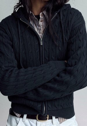 Person iført en sort kabelstrikket lynlås-sweater over en stribet skjorte med knapper, med armene over kors og hvide bukser med et sort bælte.