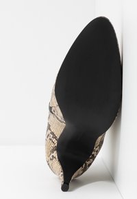 Chaussure à talon aiguilles à bout pointu avec un motif en peau de serpent beige et noir, vue par la semelle sur un fond blanc.