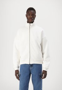 Hvit zip-up jakke med høy krage og ribbestrikkede mansjetter, laget av mykt stoff. Modellen har det på seg med blå cargo-jeans. Enkel logo på brystet.