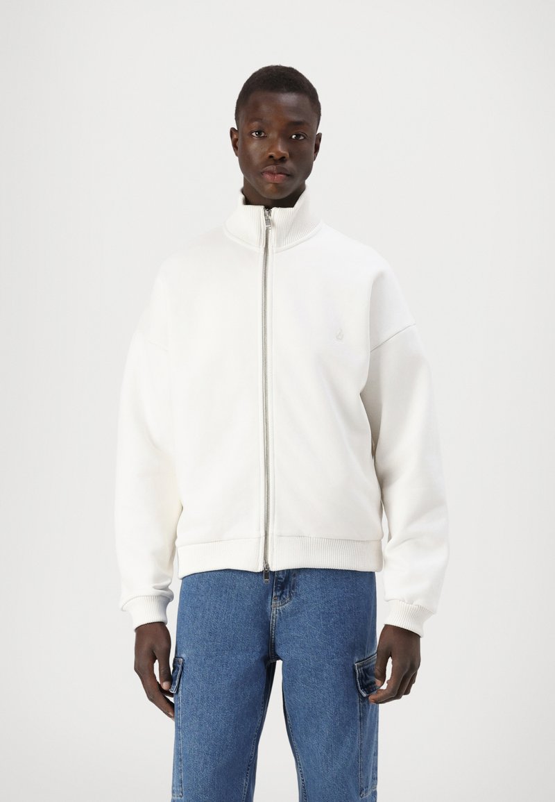 Hvit zip-up jakke med høy krage og ribbestrikkede mansjetter, laget av mykt stoff. Modellen har det på seg med blå cargo-jeans. Enkel logo på brystet.