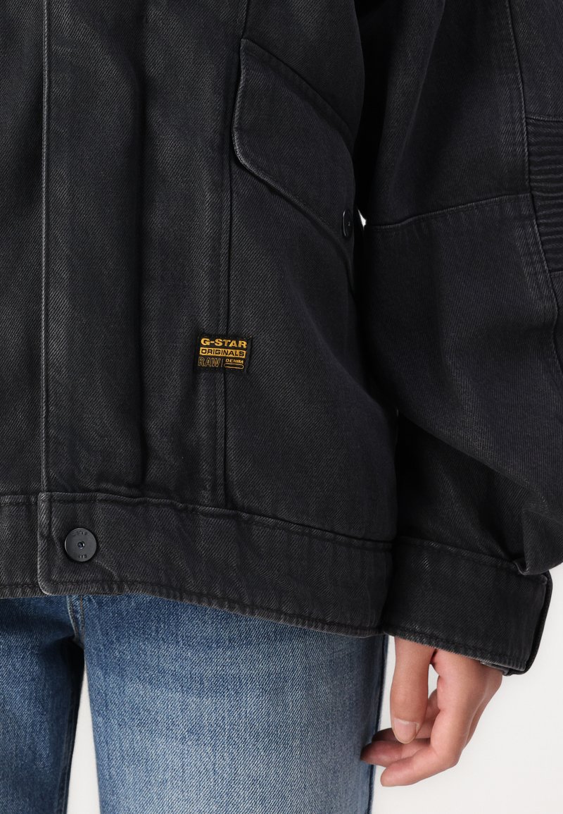 Veste en denim noir avec poche à rabat boutonnée et patch logo G-Star Originals RAW jaune, portée par-dessus un jean bleu.
