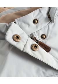 Tessuto di giacca grigio chiaro con bottoni bronzei e chiusura con zip. Accento in pelle notevole vicino alla zip. Fodera interna morbida e texturizzata visibile.
