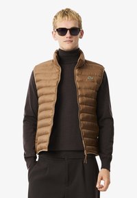Bruine gewatteerde vest met hoge kraag, ritssluiting en klein logo. Draagt over een donkere coltrui, gecombineerd met zwarte broeken.