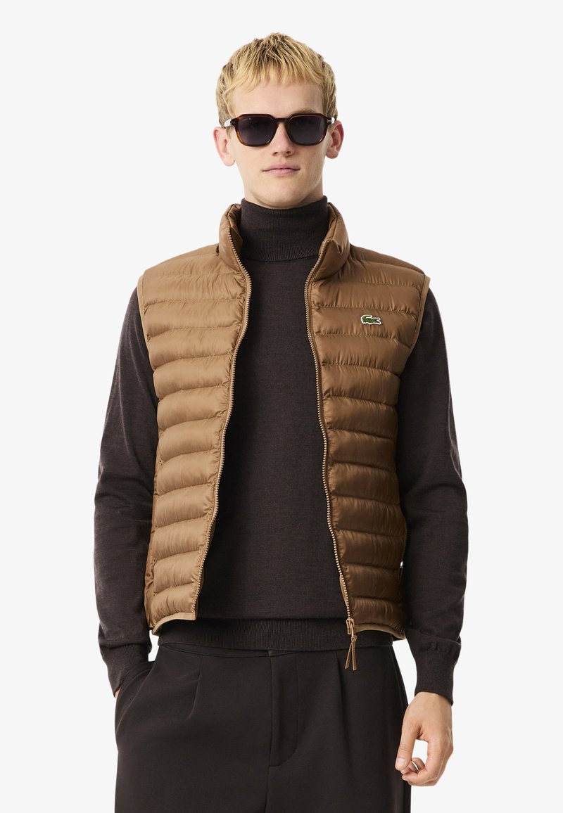 Bruine gewatteerde vest met hoge kraag, ritssluiting en klein logo. Draagt over een donkere coltrui, gecombineerd met zwarte broeken.